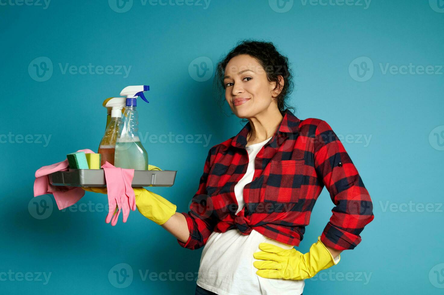 ein lockig Brünette tragen ein Weiß T-Shirt, ein rot prüfen Hemd und Gelb Gummi Handschuhe, hält ein Tablett von Reinigung liefert und süß lächelt während posieren auf ein Blau Hintergrund mit Kopieren Raum foto
