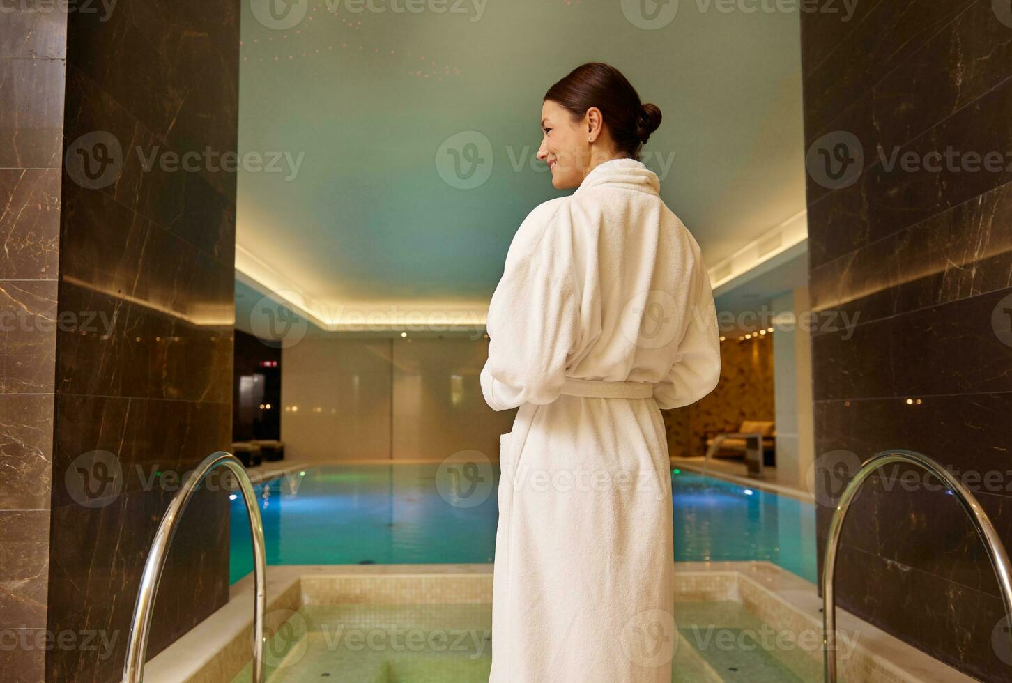 Rückseite Aussicht von ein dunkelhaarig schön Frau im Weiß Frottee Kleid Stehen durch das Schwimmbad und Whirlpool im Luxus Gesundheit Spa genießen Entspannung und Selbstpflege. Schönheit Behandlung, Entspannung, Körper Pflege Konzept foto