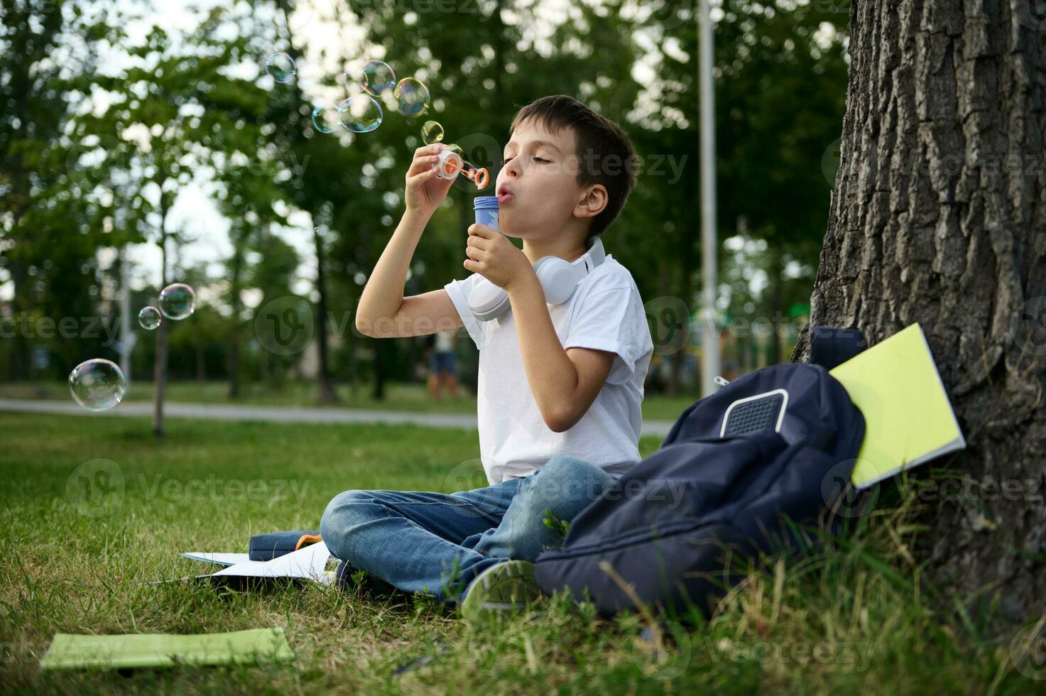 ein Schüler von elementar Noten ruhen im das Park Sitzung auf das Grün Gras nach Schule, weht Blasen. Schule Tasche mit Arbeitsmappen und Schule liefert Lügen Nieder auf das Gras foto