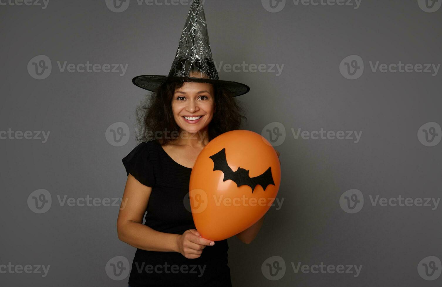 heiter jung Frau im schwarz und Magier Hut posiert mit ein Orange Luft Ballon mit ein handgemacht Filzschnitt Schläger, lächelt zahnig Lächeln suchen beim Kamera. Halloween Konzept auf grau Hintergrund, Kopieren Raum foto