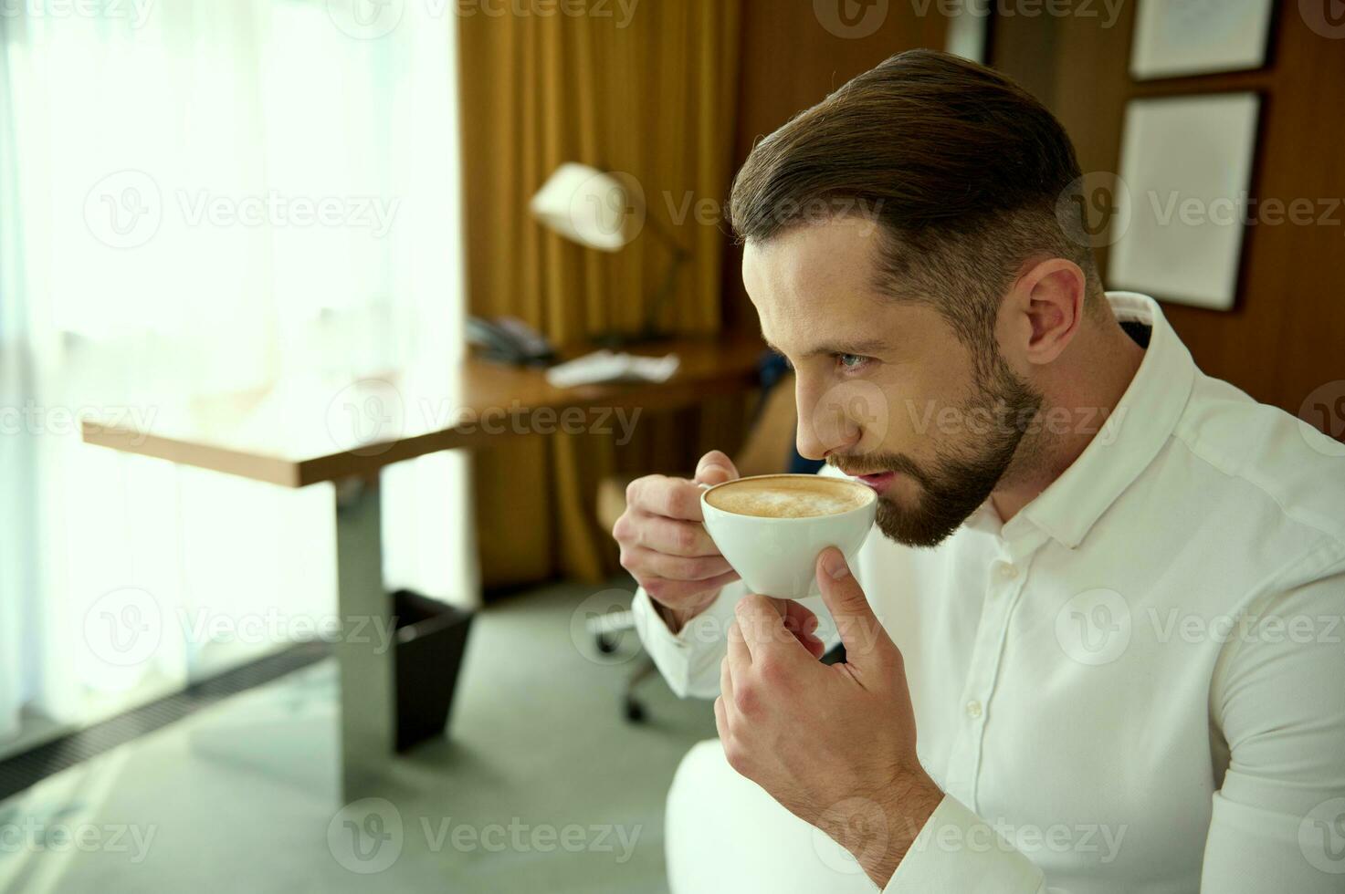 zuversichtlich attraktiv gut aussehend jung Geschäftsmann Getränke frisch gebraut Kaffee zum gut Start von das Tag, Sitzung im Hotel Zimmer während seine Geschäft Ausflug foto