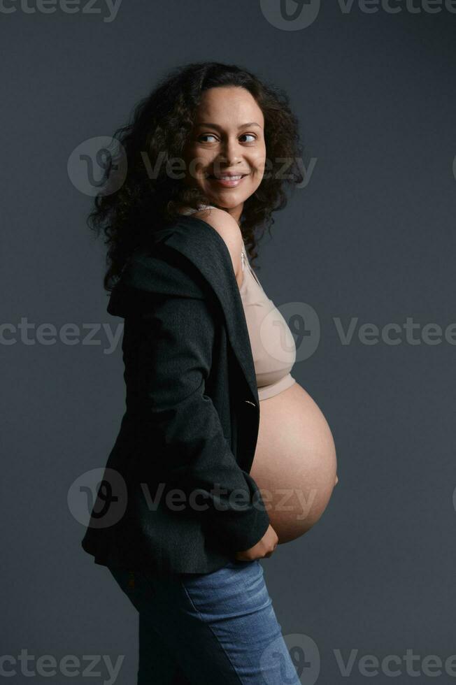 authentisch gravid Frau, halten ihr schwanger Bauch, lächelnd suchen beiseite, gekleidet im elegant Blazer, isoliert auf grau foto