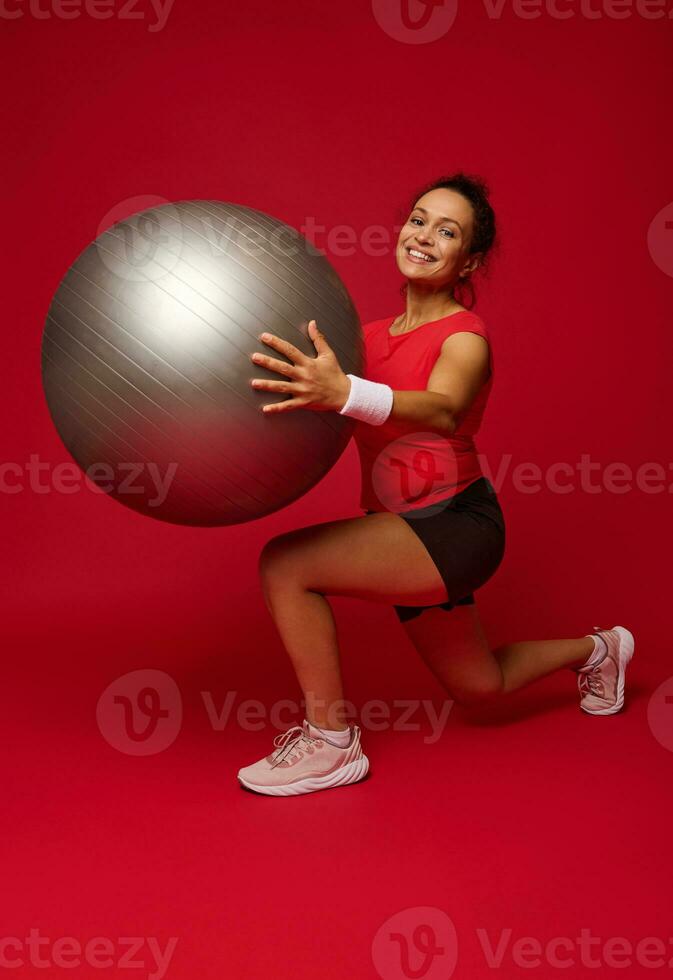 afrikanisch amerikanisch ziemlich Frau Athlet lächelt suchen beim Kamera während durchführen nach vorne Ausfallschritte mit passen Ball gegen rot farbig Hintergrund mit Kopieren Anzeige Raum foto