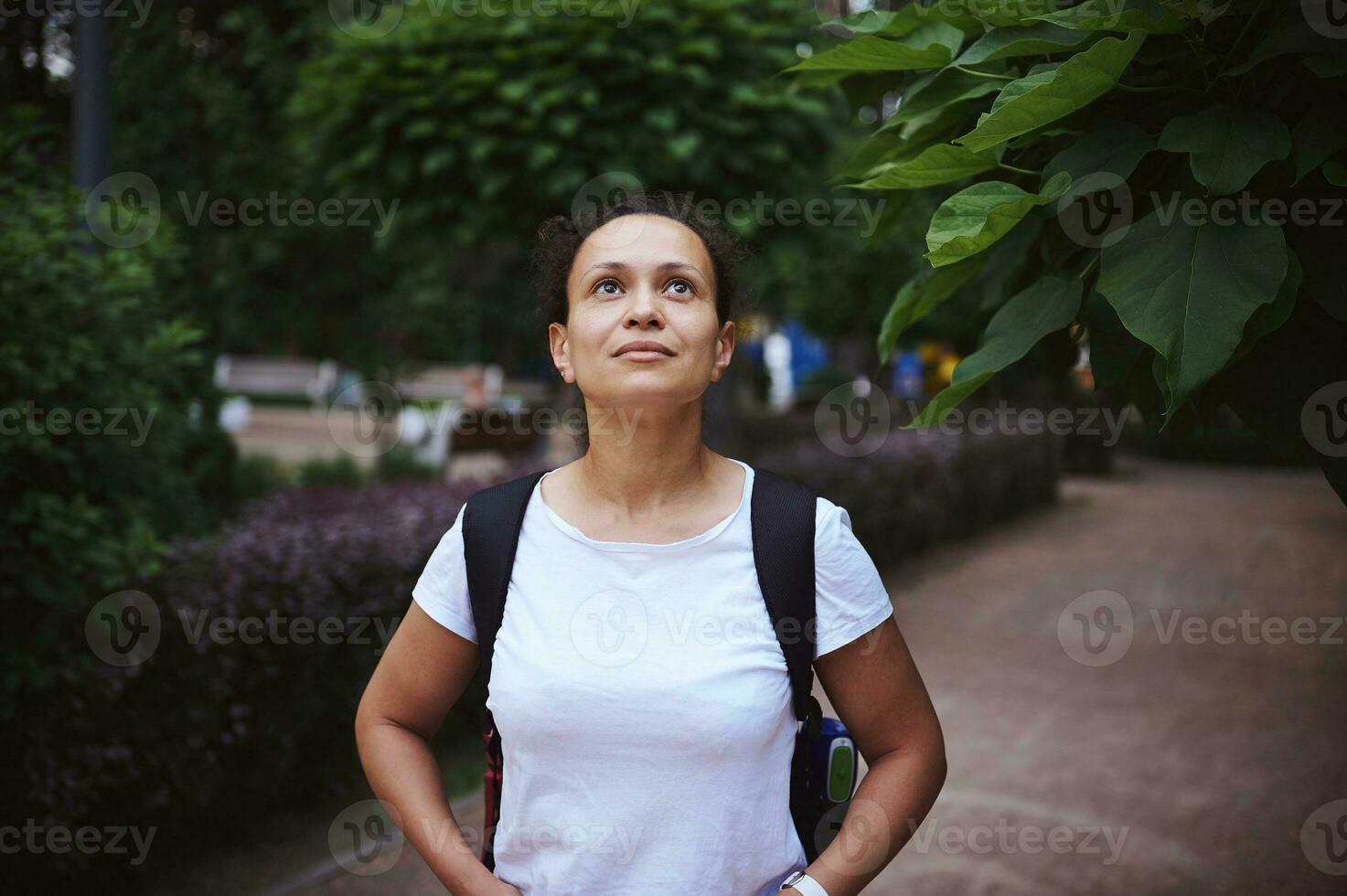 zuversichtlich entspannt multiethnisch Frau Tourist mit Rucksack Gehen entlang das Gasse im das Park auf ein Sommer- warm Tag foto