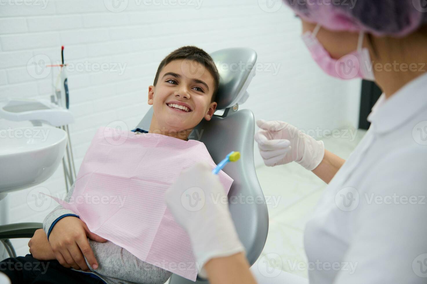 bezaubernd Junge Sitzung im Zahnarzt Stuhl lächelt mit schön zahnig Lächeln suchen beim ein pädiatrisch Zahnarzt Lehren ihm Über Oral Hygiene während Dental geplanter Termin im Weiß sauber modern medizinisch Kabinett foto