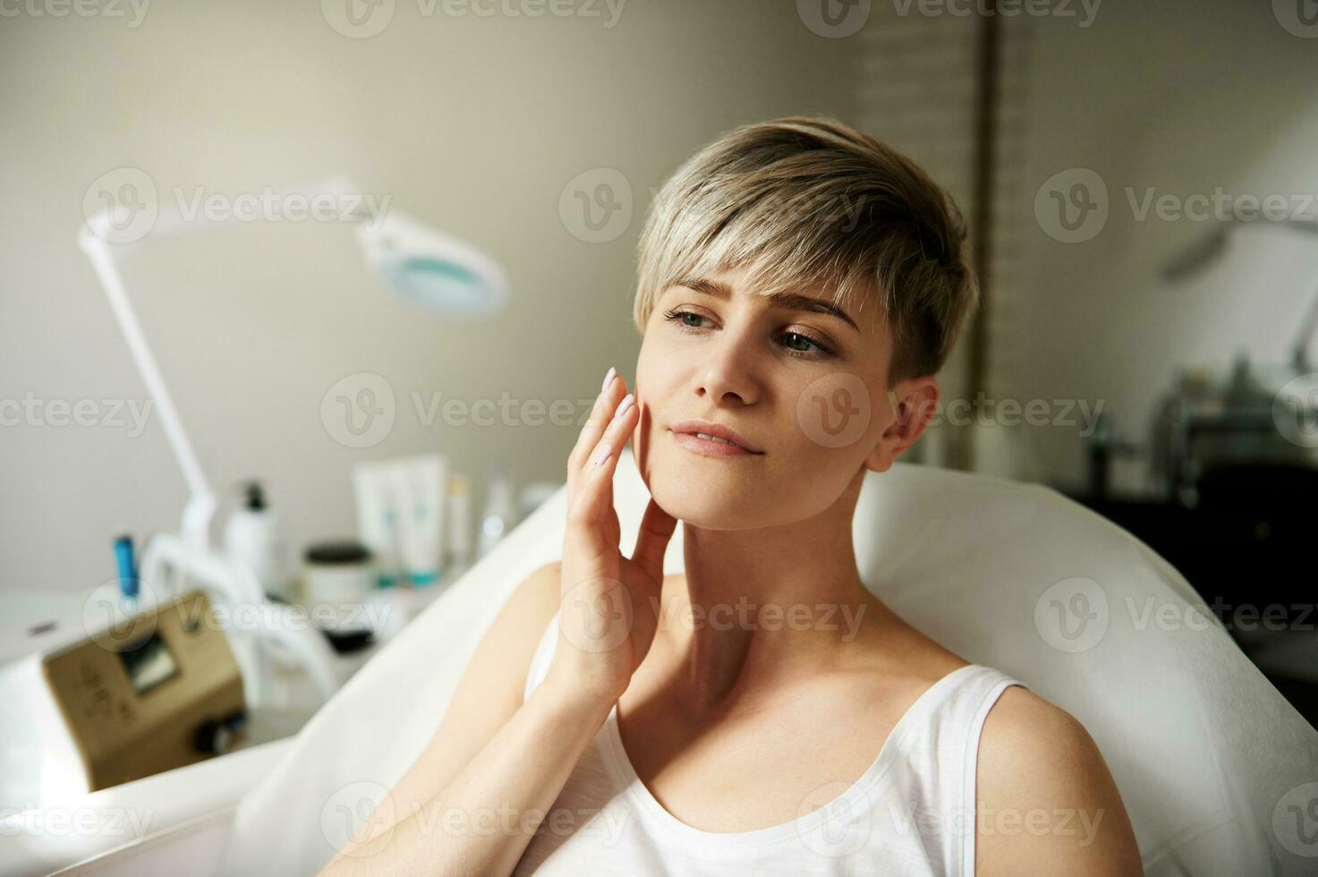 schön jung blond mit kurz Haar sanft berühren ihr Gesicht während Sitzung auf ein Stuhl im ein Schönheit Salon im ein Spa Klinik foto