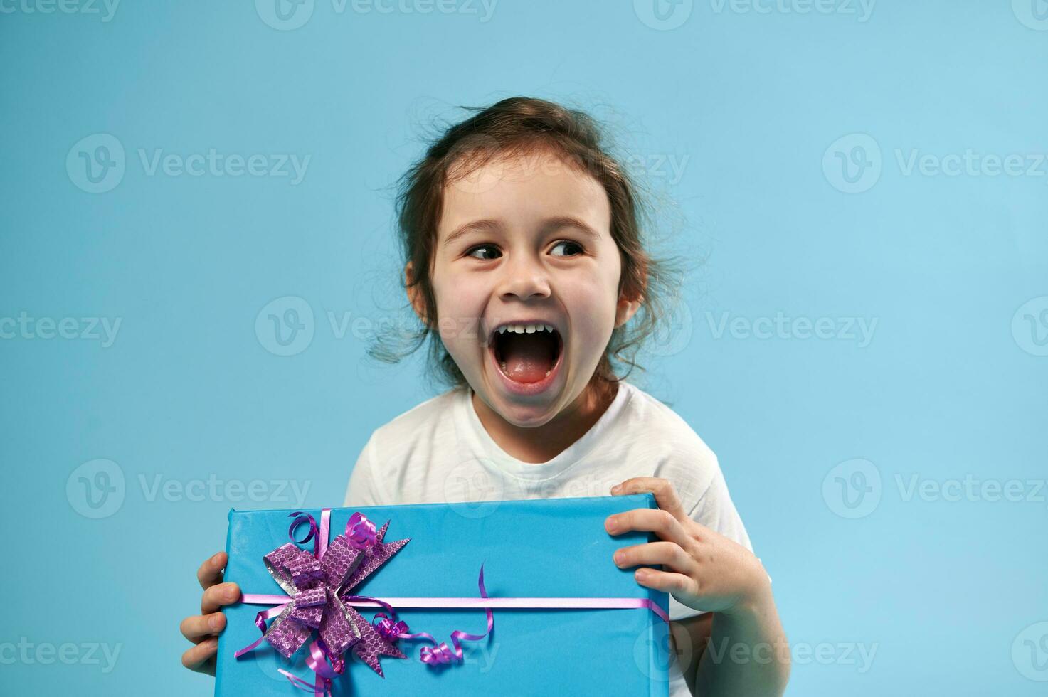 süß wenig Mädchen Lachen während posieren auf Blau Hintergrund mit Geschenk im ihr Hände. Kopieren Raum. Feier Konzept. foto