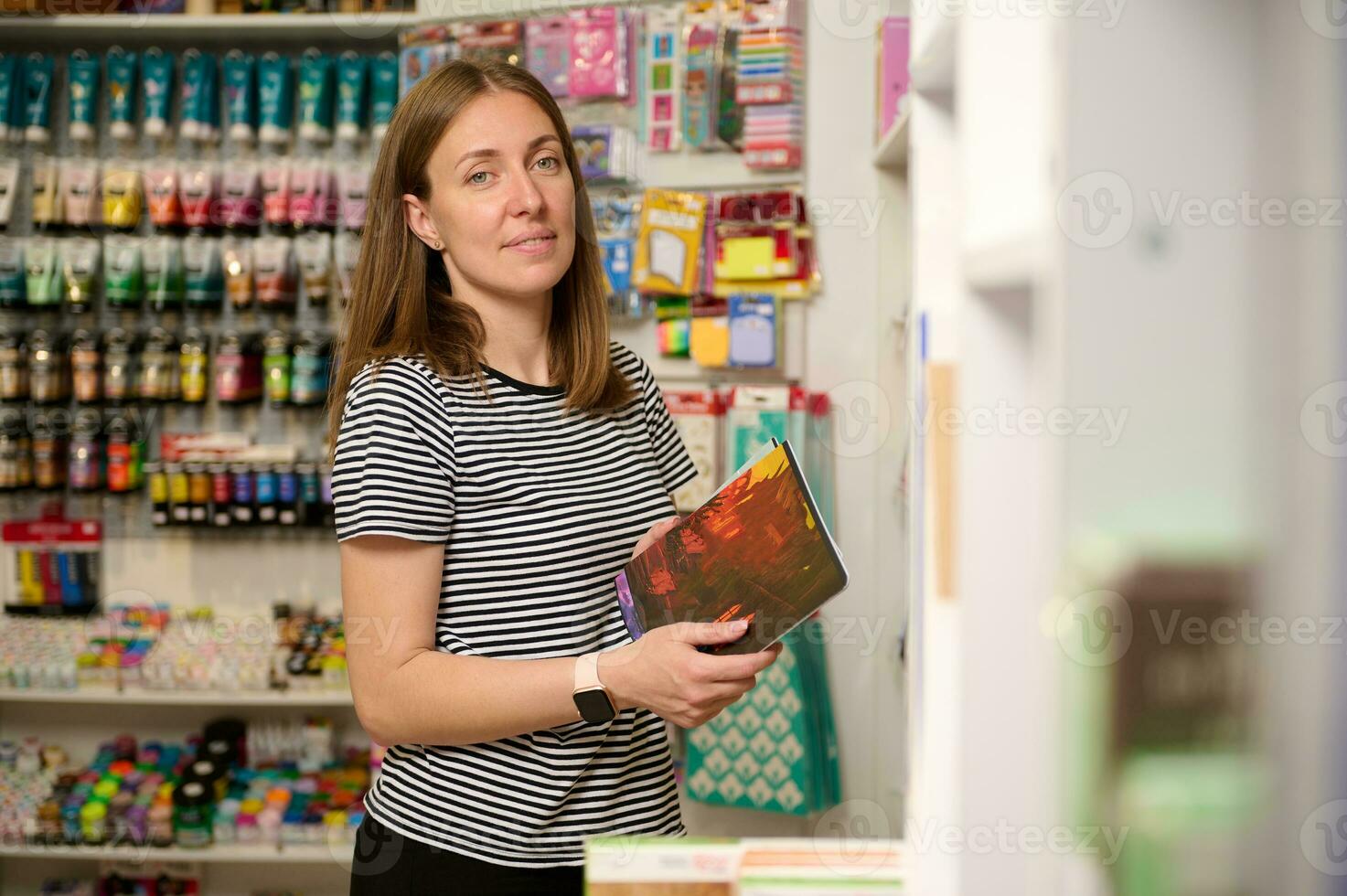 angenehm weiblich Verkäufer hält ein Notizbuch, lächelt suchen beim Kamera während funktioniert auf Merchandising im Schule Schreibwaren Geschäft foto