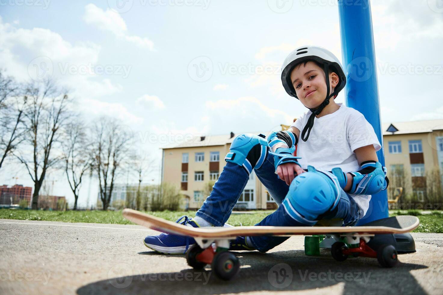 Unterseite Aussicht von lächelnd Junge im schützend Ausrüstung von Skateboardfahrer Sitzung auf Skateboard und suchen beim Kamera foto