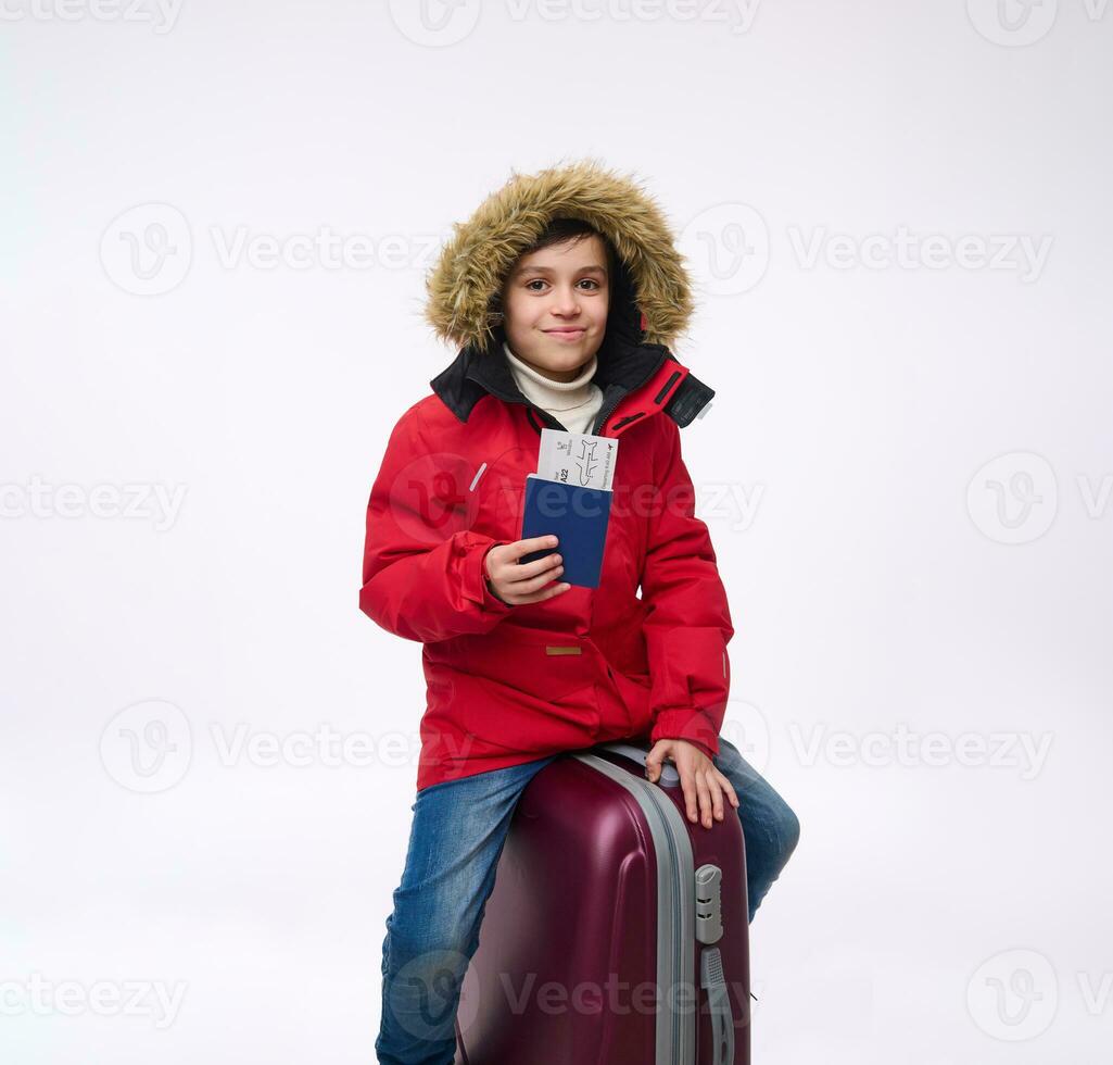 heiter jugendlich, elementar alt Junge im hell rot Nieder Jacke Sitzung auf seine Gepäck, suchen beim Kamera und zeigen Reisepass mit Einsteigen passieren. Winter Feiertage, Reise Konzept mit Kopieren Anzeige Raum foto
