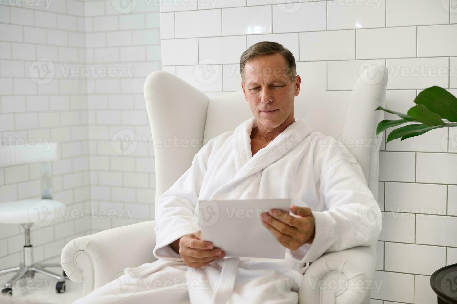 gut aussehend reifen europäisch Mann gekleidet im Weiß Bademantel, mit ein leer Papier Blatt, Sitzung Sessel im luxuriös Wellness Spa Center foto