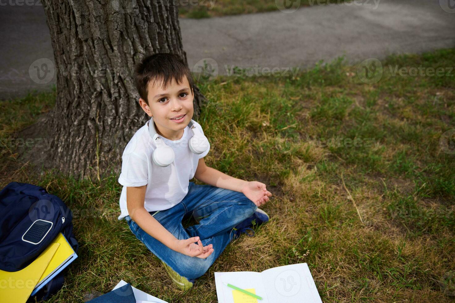 gut aussehend Schule Kind Junge Sitzung im Lotus Position auf das Grün Gras von Öffentlichkeit Park und meditieren , suchen oben beim das Kamera, süß lächelnd. Arbeitsmappen und Schule liefert Lügen auf das Gras foto