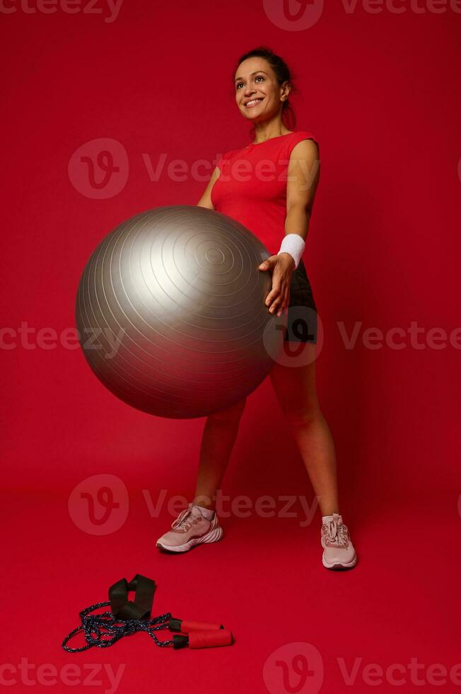voll Länge Porträt von ein schön aktiv Frau im Sportbekleidung ausüben mit passen Ball gegen rot farbig Hintergrund mit Kopieren Raum foto
