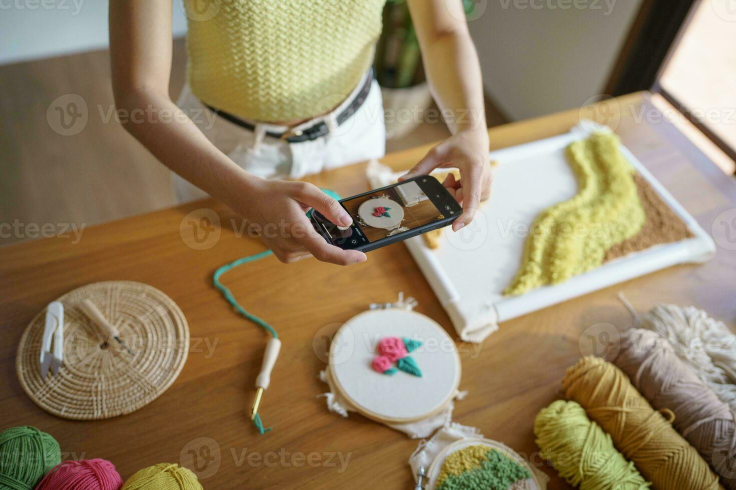 asiatisch Frau nehmen Smartphone Foto schlagen Nadel. Telefon Buchung auf Sozial Netzwerke im Studio Werkstatt. Designer Arbeitsplatz handgemacht Kunst Projekt DIY Stickerei