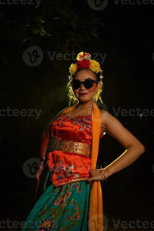 ein Sundanesisch Tänzer posiert im Vorderseite von ein leuchtenden Licht während tragen ein Orange Kostüm mit Sonnenbrille und Blumen auf ihr Kopf foto