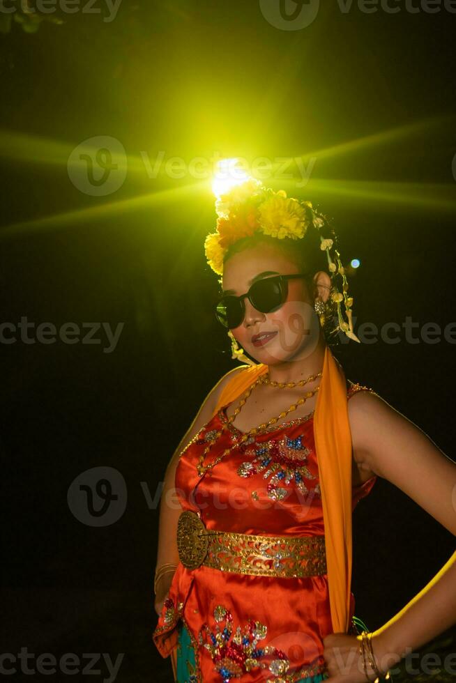 ein Sundanesisch Tänzer posiert im Vorderseite von ein leuchtenden Licht während tragen ein Orange Kostüm mit Sonnenbrille und Blumen auf ihr Kopf foto