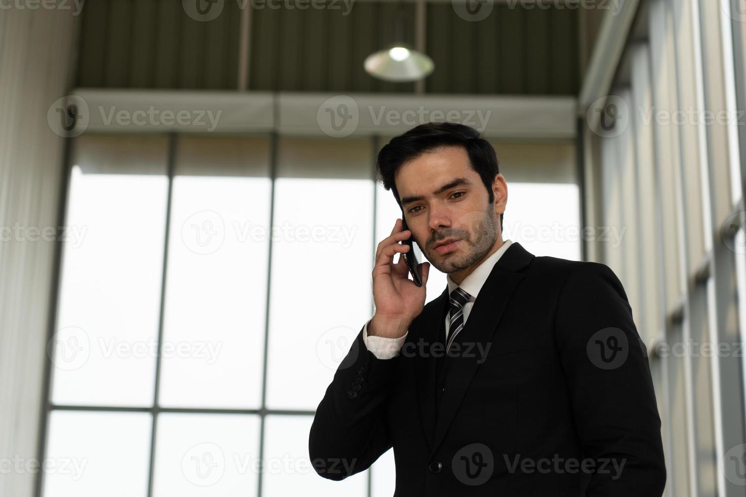 Offenes Porträt eines jungen, gutaussehenden, kaukasischen, selbstbewussten Geschäftsmannes im schwarzen Anzug mit Krawatte, der das Smartphone verwendet, um mit dem Kunden im Büro zu sprechen? foto