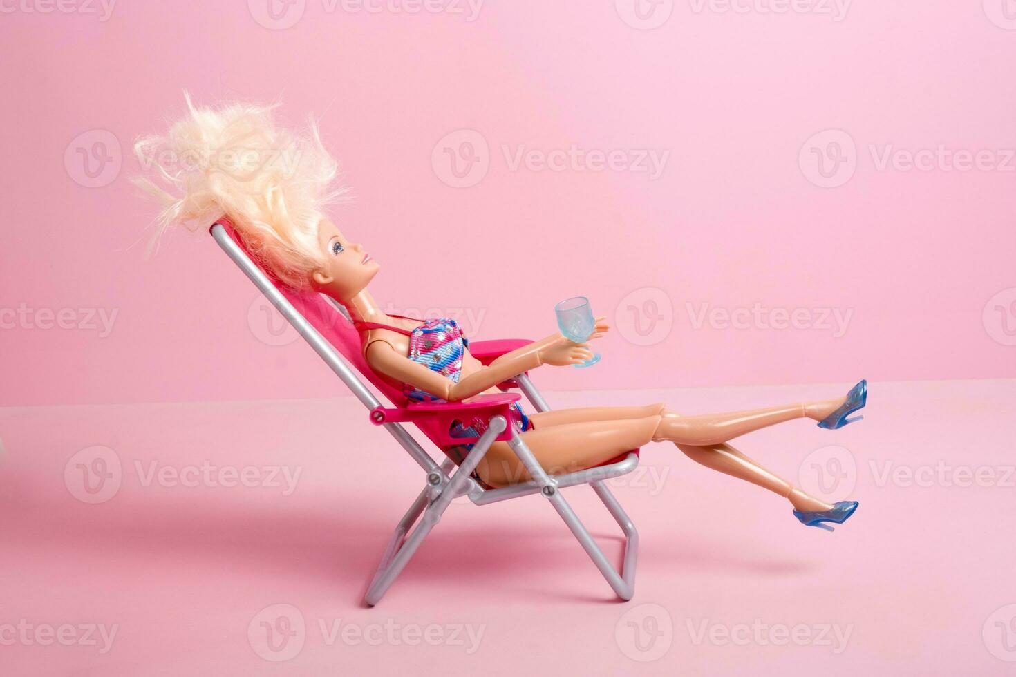 ein Puppe im ein Badeanzug mit ein Glas auf das Strand auf ein Rosa Hintergrund. Strand Ferien Konzept foto