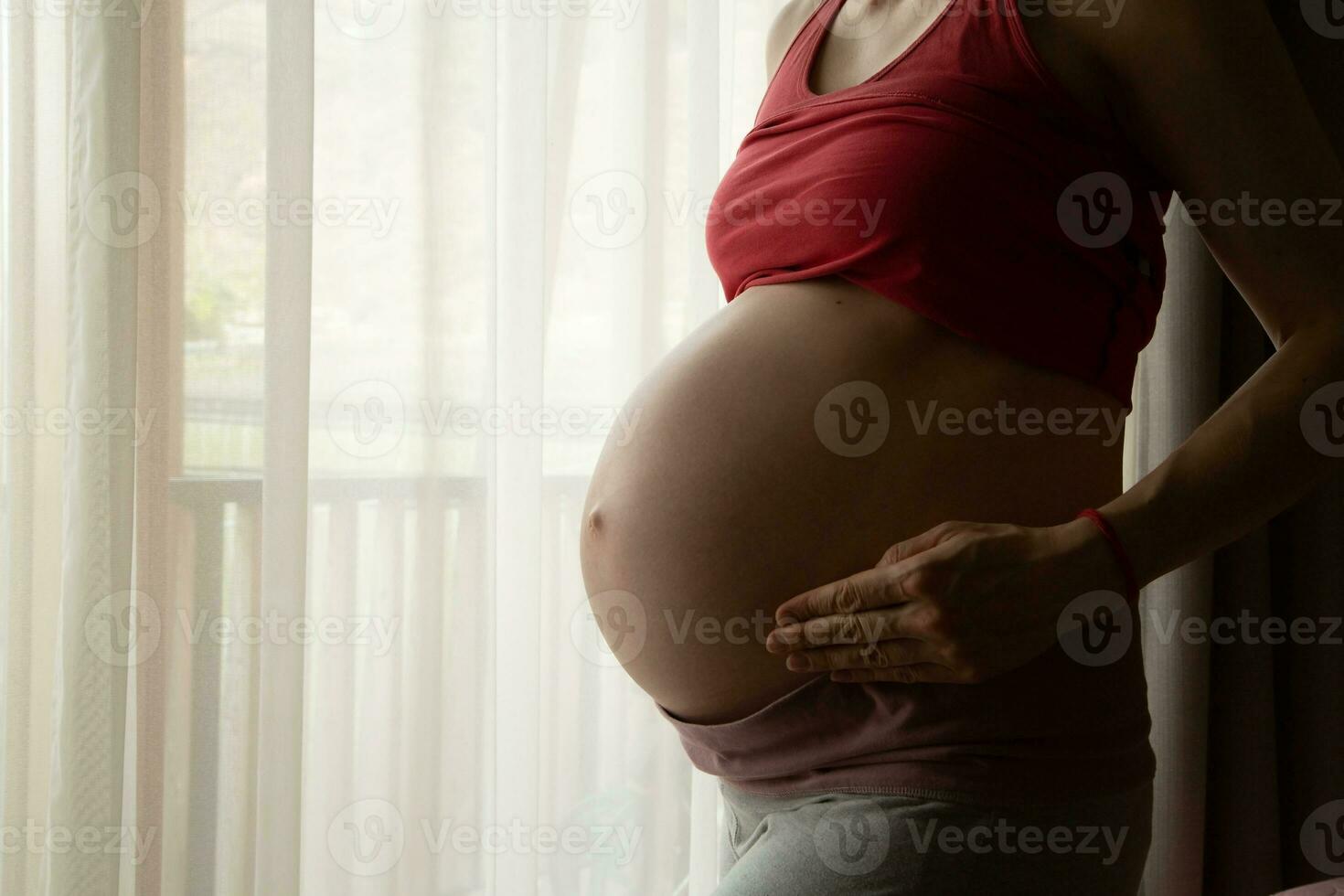 ein schwanger Frau im ein rot oben ist Stehen im Vorderseite von ein Fenster foto