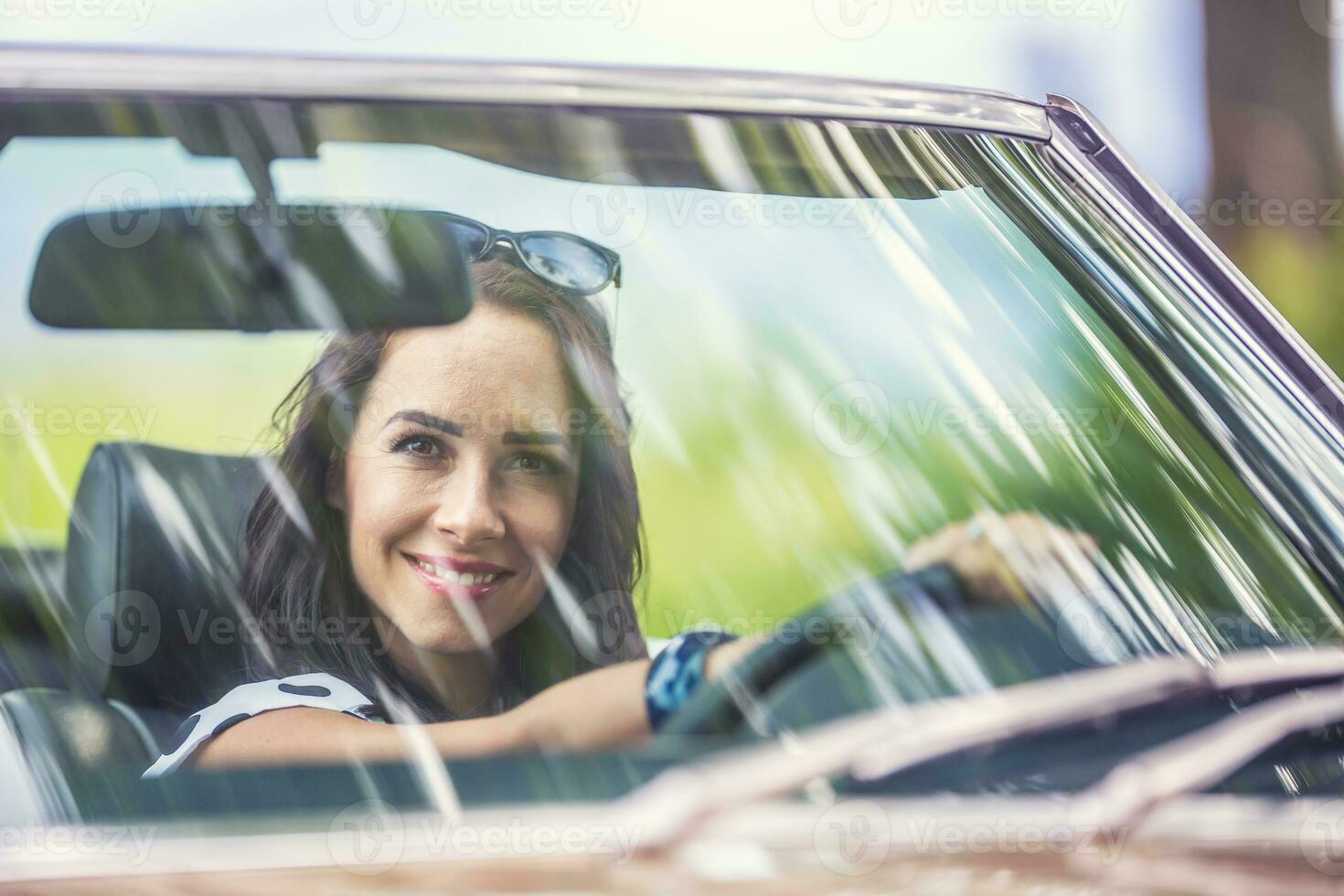 linke Hand Fahren weiblich sitzt hinter das Lenkung Rad von ein Auto, lächelnd in das Kamera durch Vorderseite Fenster foto