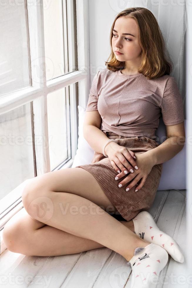 Nachdenkliches schönes junges Mädchen sitzt auf einem Fensterbrett am Fenster foto