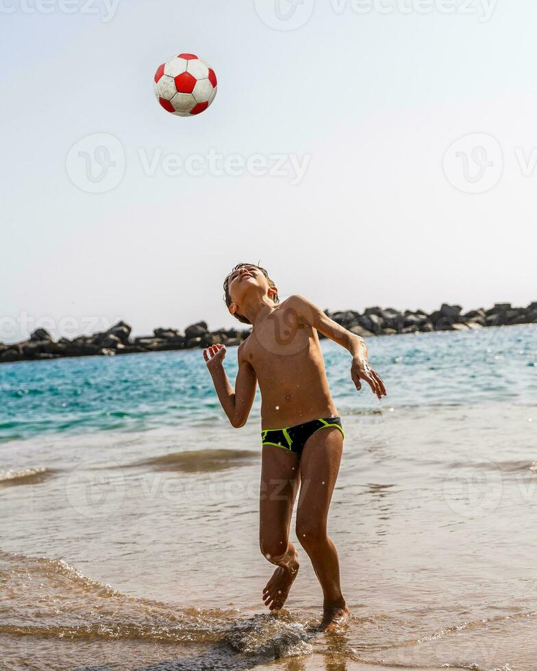 jung Junge spielen mit ein Fußball Ball auf das Strand durch das Meer foto