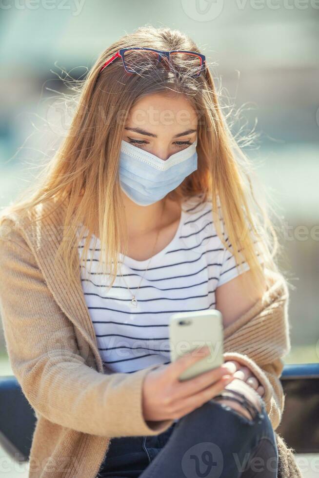 Jugend Mädchen Sitzung draußen SMS auf ein Handy tragen schützend Gesicht Maske foto