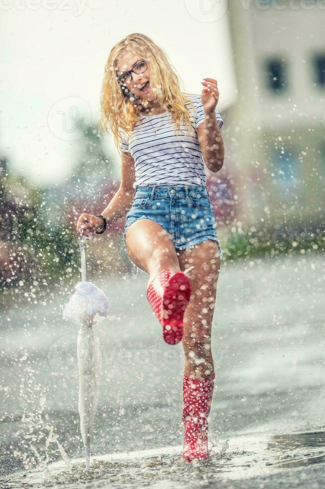 heiter Mädchen Springen mit Weiß Regenschirm im gepunktet rot Galoschen. heiß Sommer- Tag nach das Regen Frau Springen und planschen im Pfütze foto