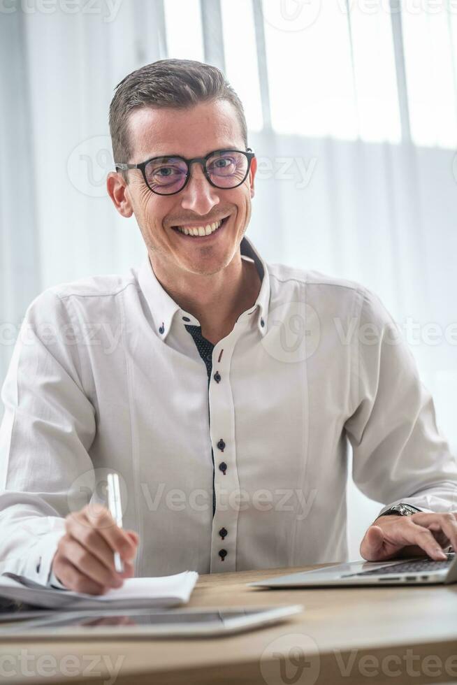 jung Geschäftsmann tragen Brille lächelt beim das Kamera während Sitzung beim seine Büro Schreibtisch foto