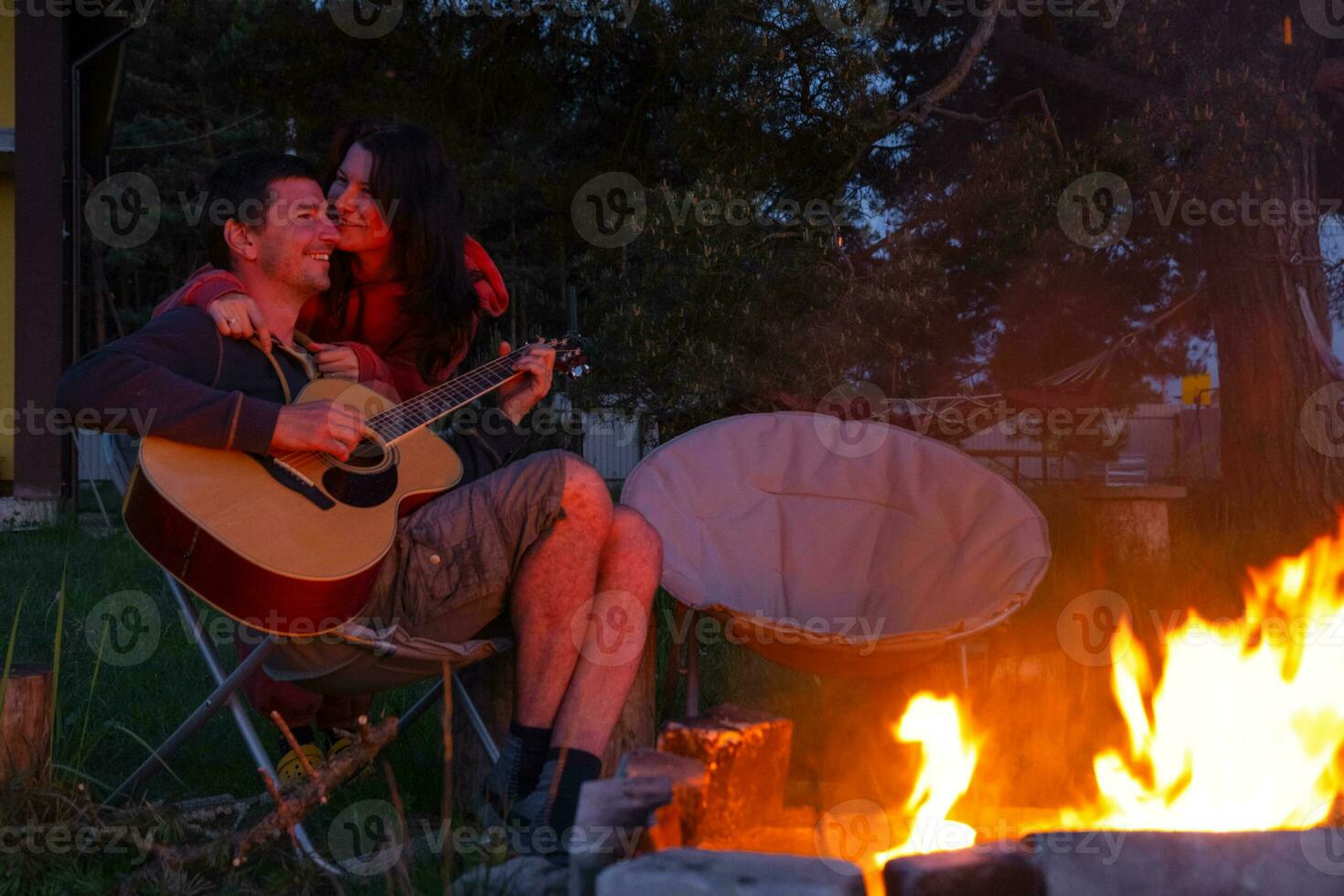 ein Mann Theaterstücke das Gitarre, ein Frau hört zu und singt entlang. ein Paar im Liebe ist Sitzung durch das draussen Lagerfeuer im das Hof von das Haus auf Camping Stühle, ein romantisch Abend foto
