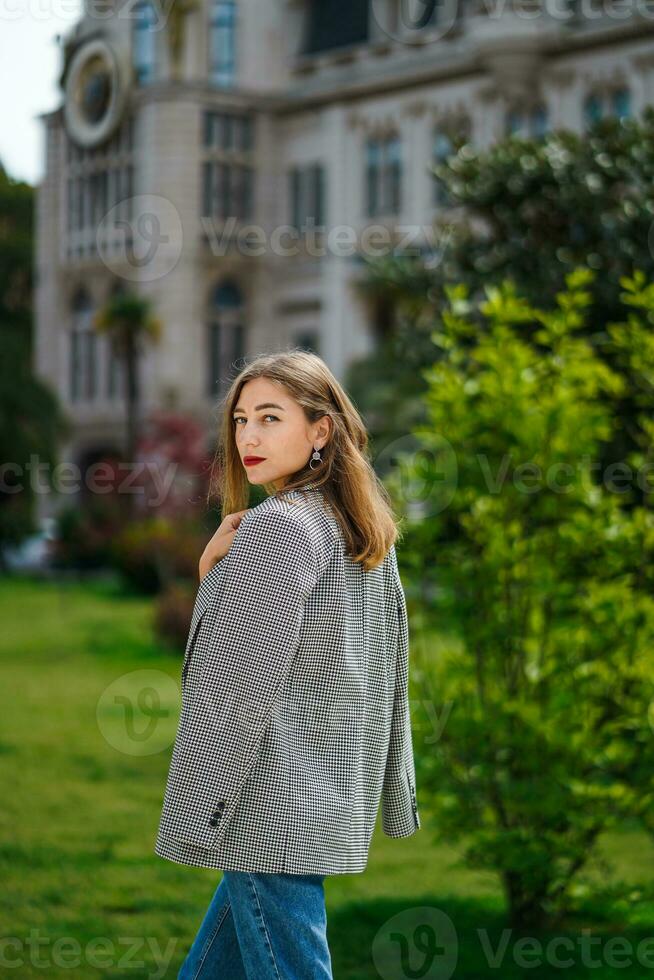 Mädchen auf Stadt gehen. jung Frau Stehen mit ihr zurück im Hälfte Wende und suchen direkt beim Kamera. cool Wetter im Sommer, Mädchen drapiert Jacke Über ihr Schultern. alt Gebäude und Bäume auf lange planen. foto