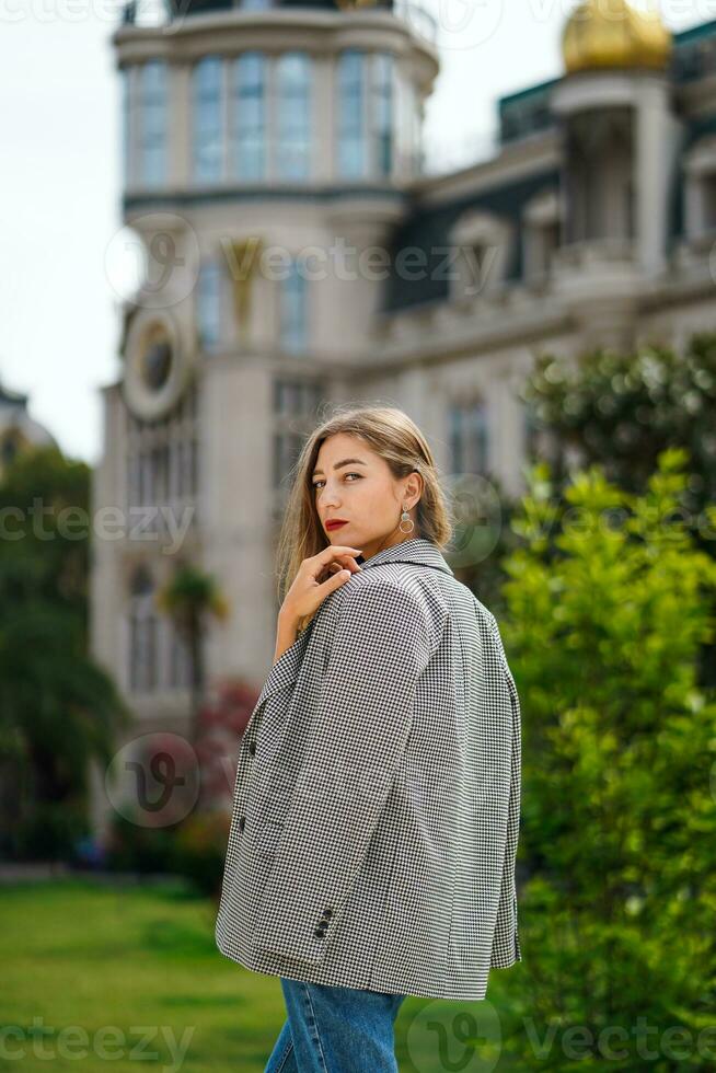 Frau Stehen auf Straße. schön jung Frau tragen Jacke und Jeans gehen. Grün Bäume und historisch Gebäude Fassade auf Hintergrund. Stadt Erholung. sorglos Person ruhen erleben Ruhe foto