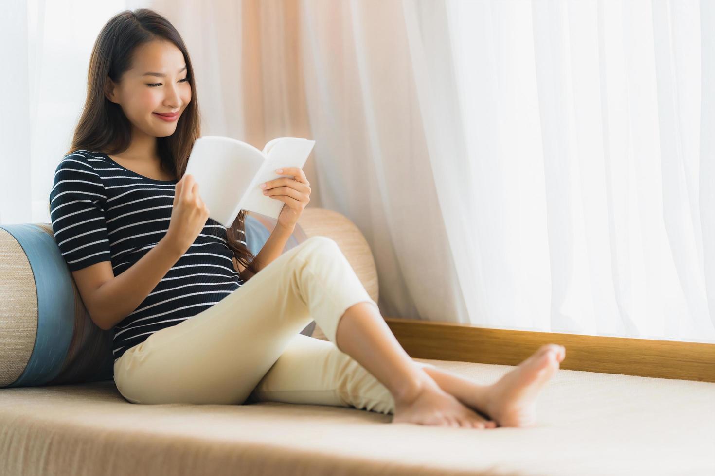 Porträt der schönen jungen Asiatin, die ein Buch auf dem Sofa im Wohnzimmer liest foto