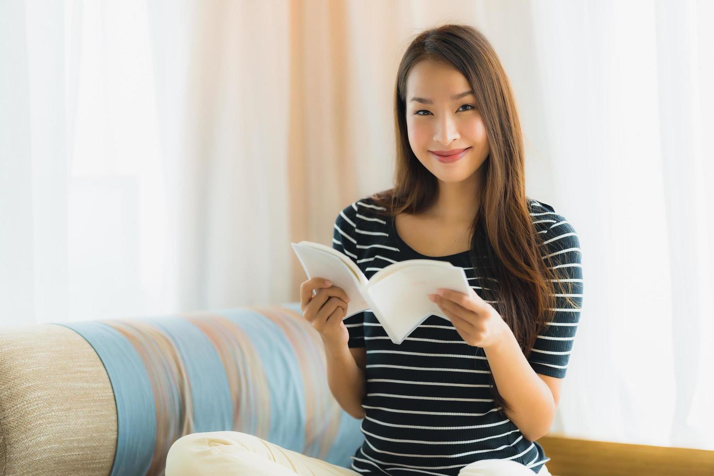 Porträt der schönen jungen Asiatin, die ein Buch auf dem Sofa im Wohnzimmer liest foto