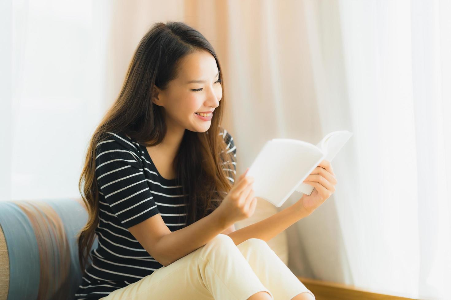 Porträt der schönen jungen Asiatin, die ein Buch auf dem Sofa im Wohnzimmer liest foto