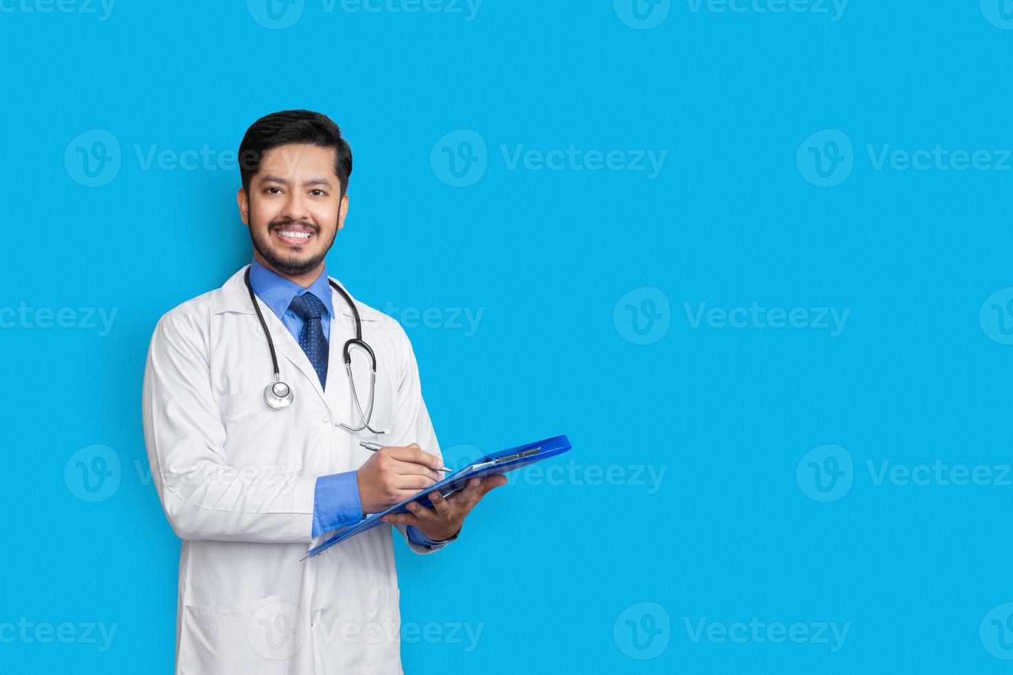 Arzt im Laborkittel hält Patientenakte oder schreibt Krankenakte mit Blick in die Kamera, isoliert auf blauem Hintergrund foto