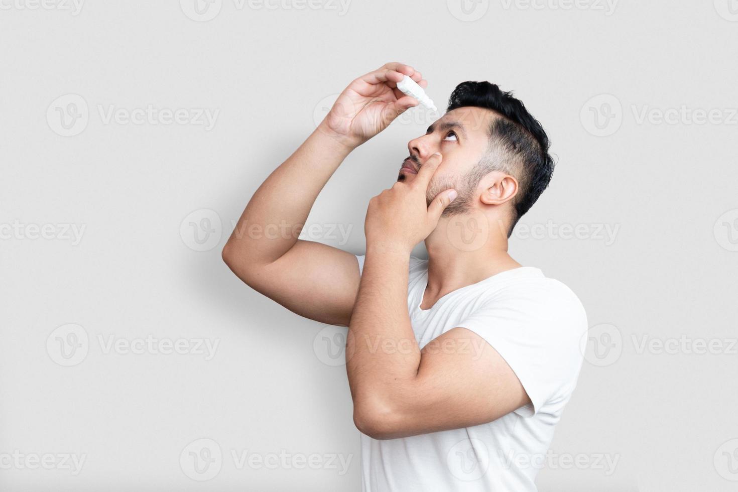 Vision und Augenheilkunde, Gesundheitskonzept, Seitenansichtporträt eines gutaussehenden jungen Mannes in weißem T-Shirt, der Augentropfen auf weißem Hintergrund aufträgt foto