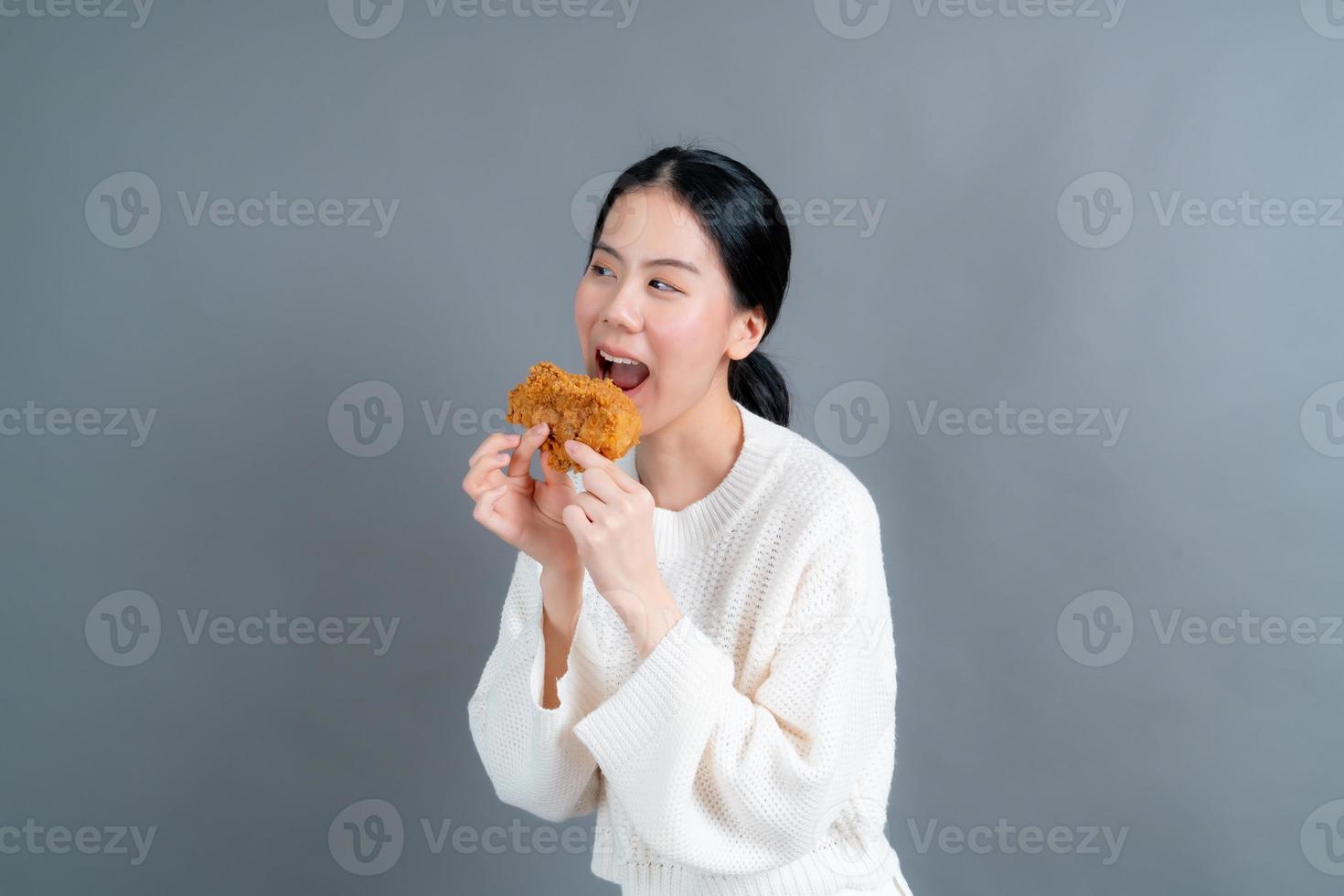 junge asiatische frau trägt einen pullover mit einem glücklichen gesicht und genießt es, gebratenes hähnchen auf grauem hintergrund zu essen foto