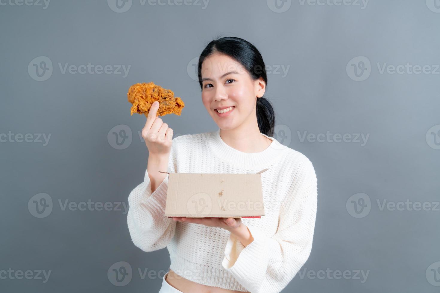 junge asiatische frau trägt einen pullover mit einem glücklichen gesicht und genießt es, gebratenes hähnchen auf grauem hintergrund zu essen foto