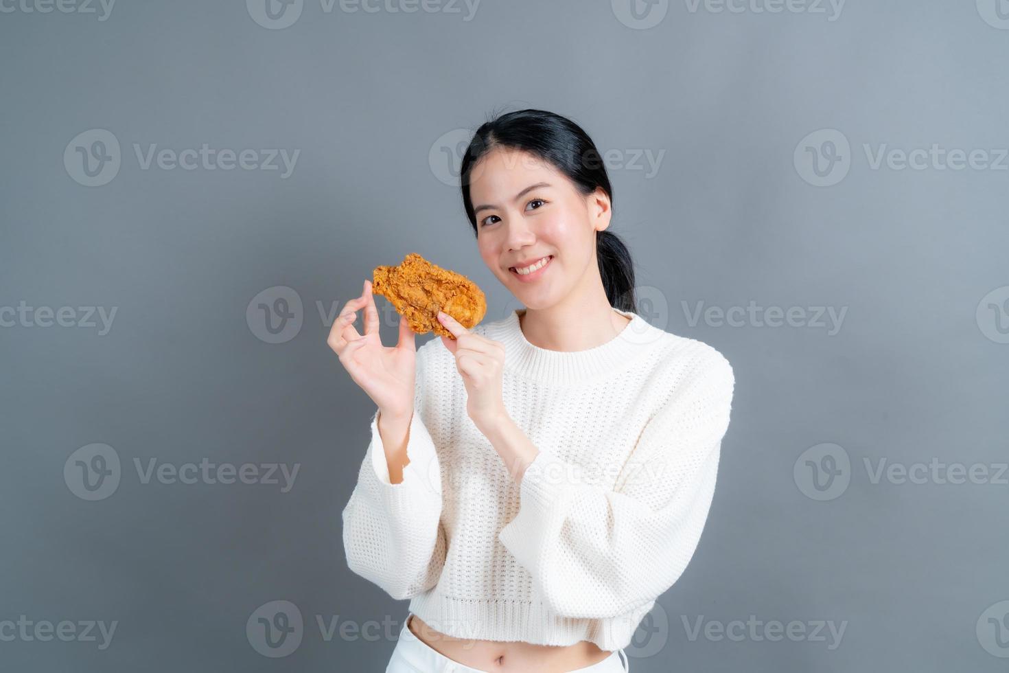 junge asiatische frau trägt einen pullover mit einem glücklichen gesicht und genießt es, gebratenes hähnchen auf grauem hintergrund zu essen foto