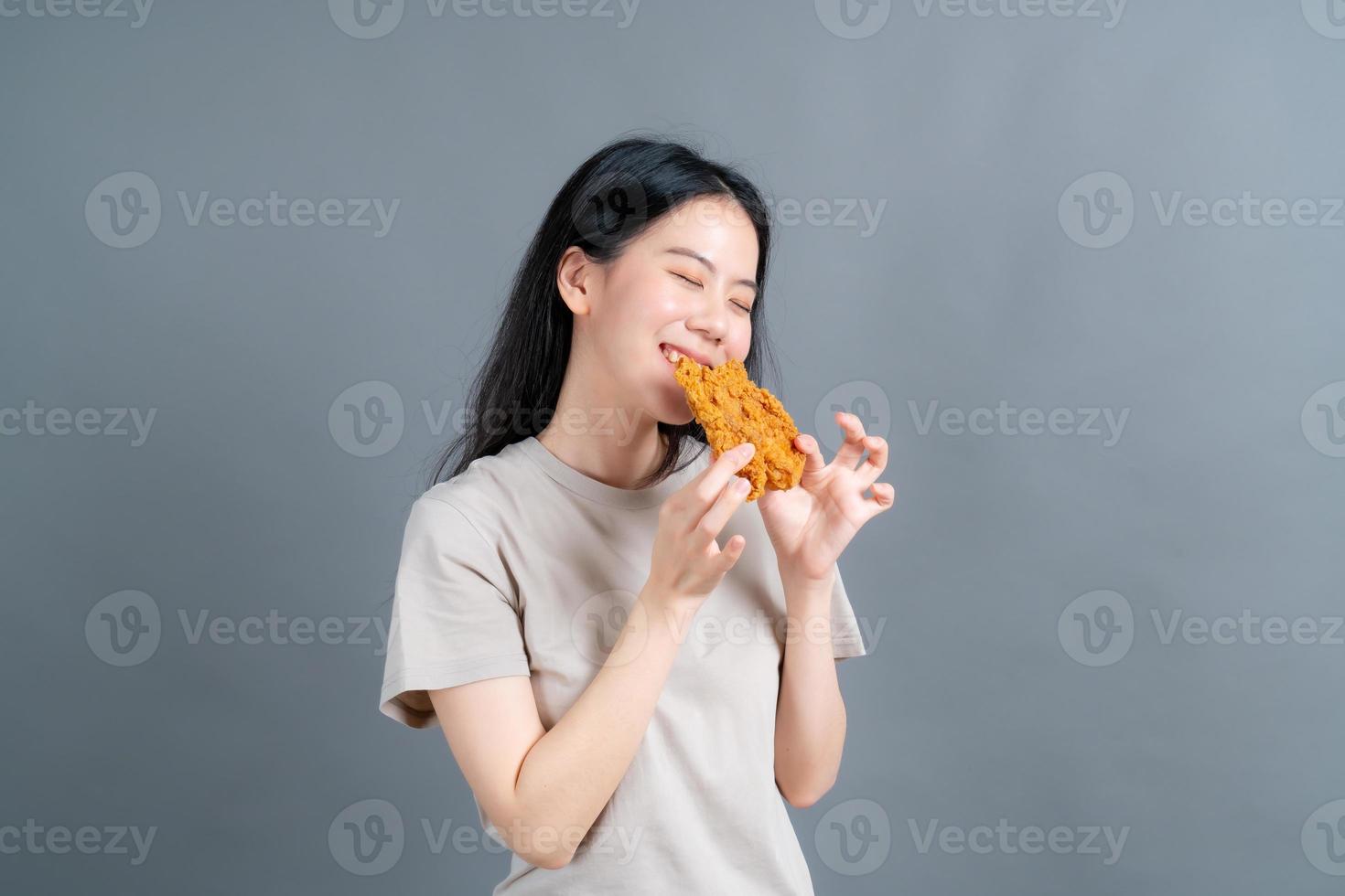 junge asiatische frau trägt einen pullover mit einem glücklichen gesicht und genießt es, gebratenes hähnchen auf grauem hintergrund zu essen foto