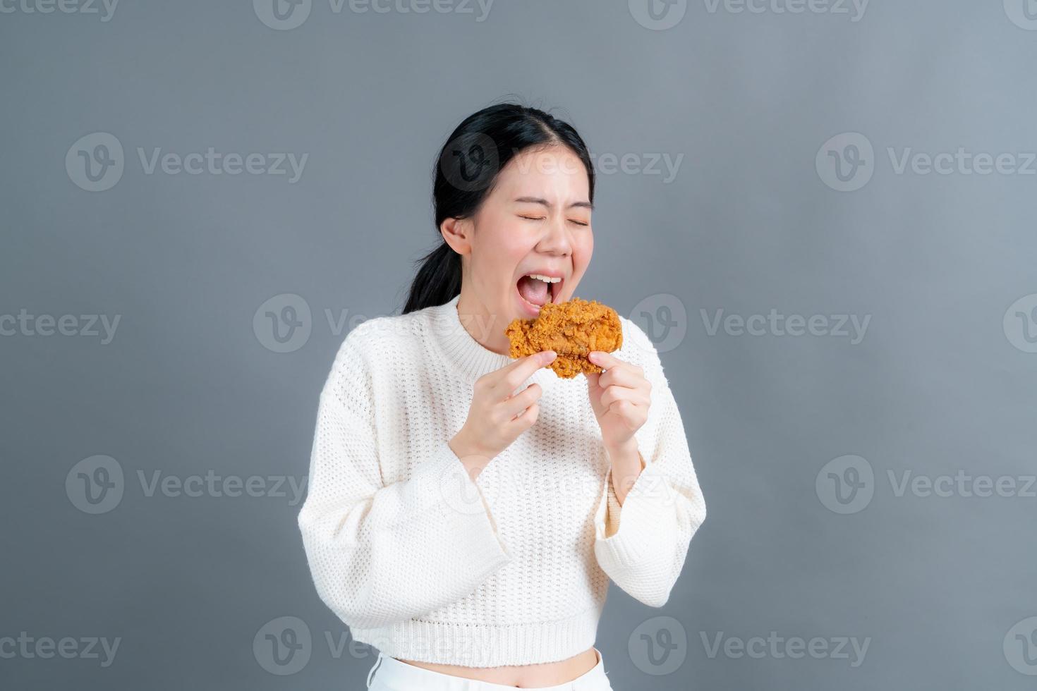 junge asiatische frau trägt einen pullover mit einem glücklichen gesicht und genießt es, gebratenes hähnchen auf grauem hintergrund zu essen foto