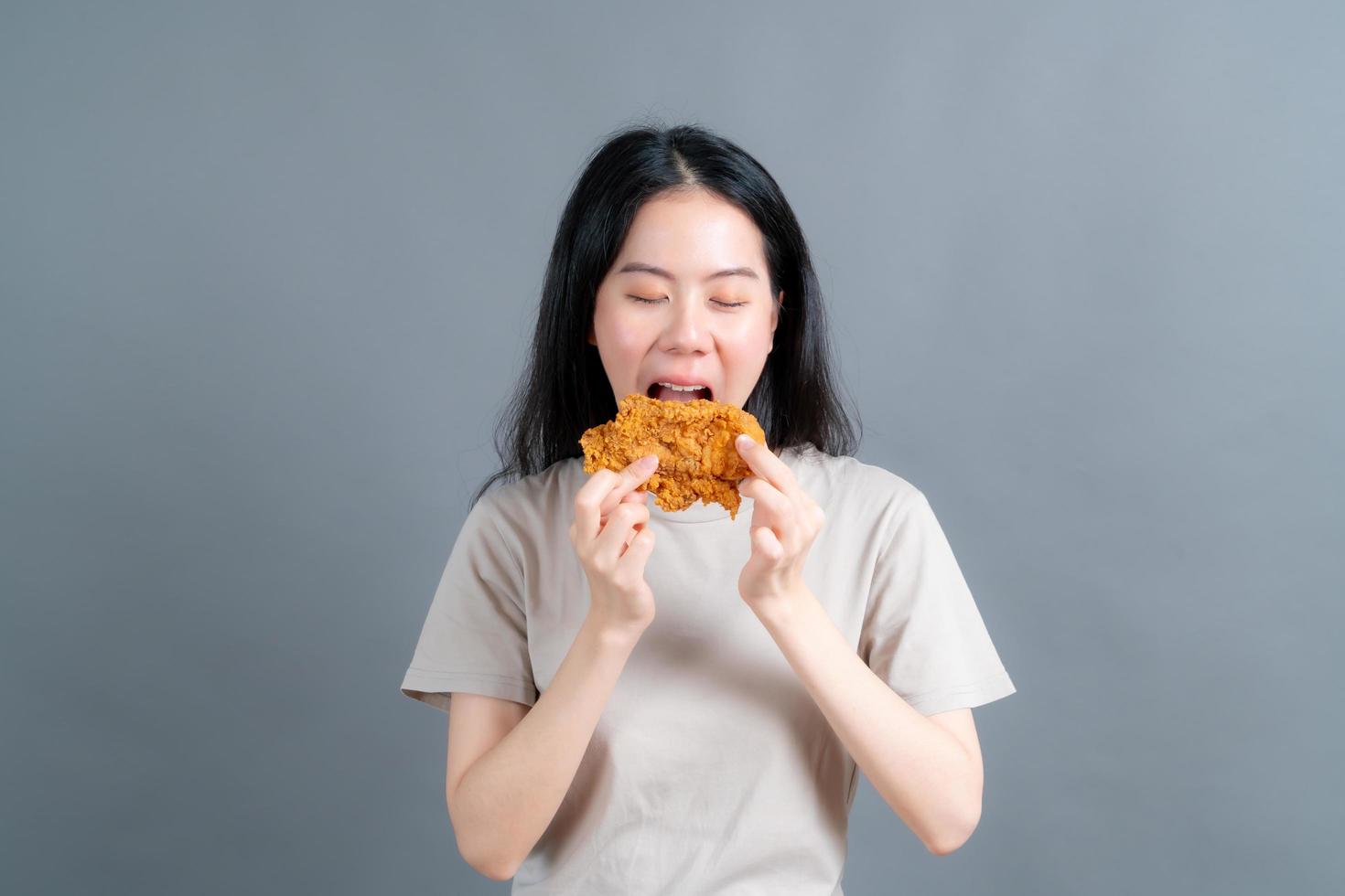 junge asiatische frau trägt einen pullover mit einem glücklichen gesicht und genießt es, gebratenes hähnchen auf grauem hintergrund zu essen foto