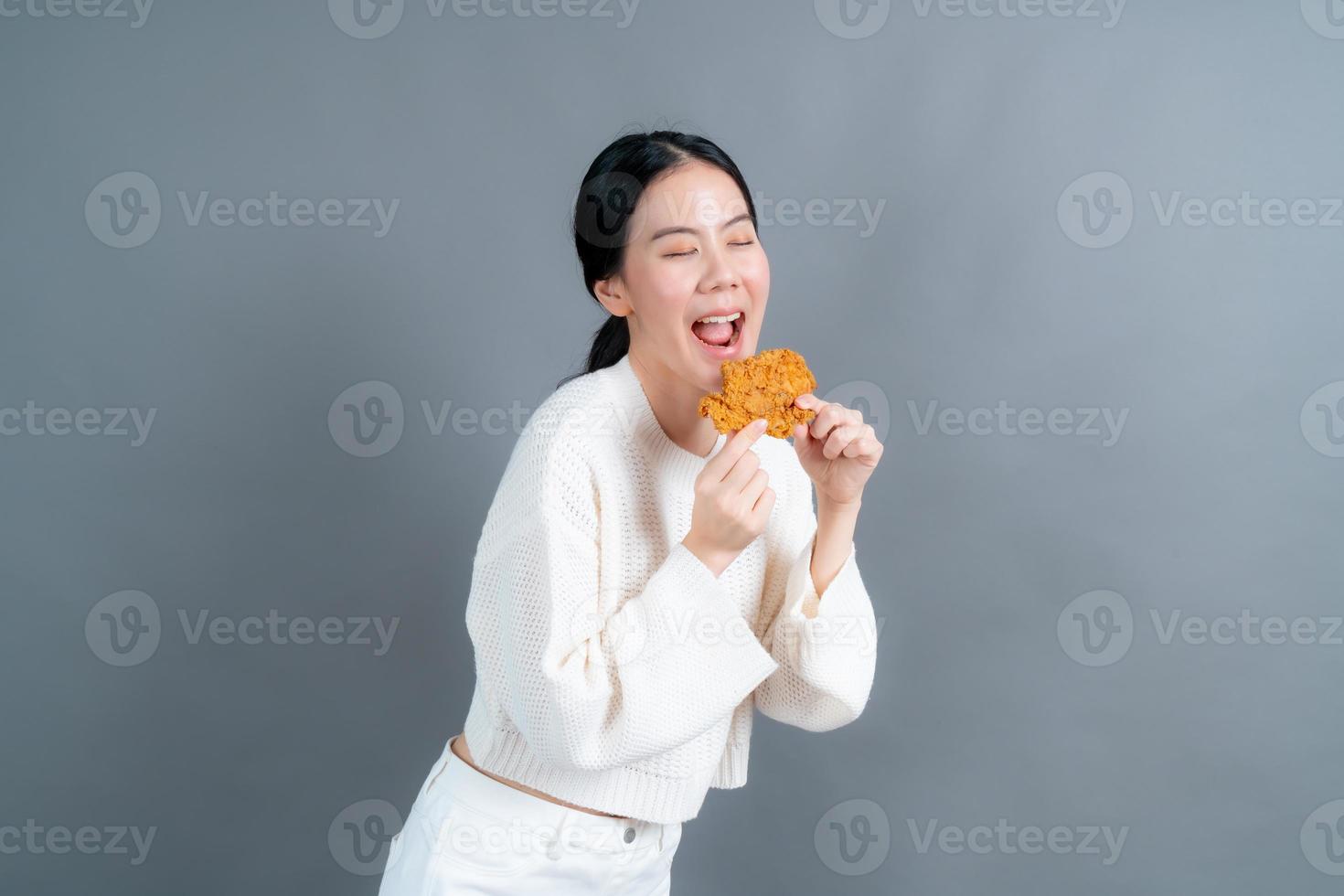 junge asiatische frau trägt einen pullover mit einem glücklichen gesicht und genießt es, gebratenes hähnchen auf grauem hintergrund zu essen foto