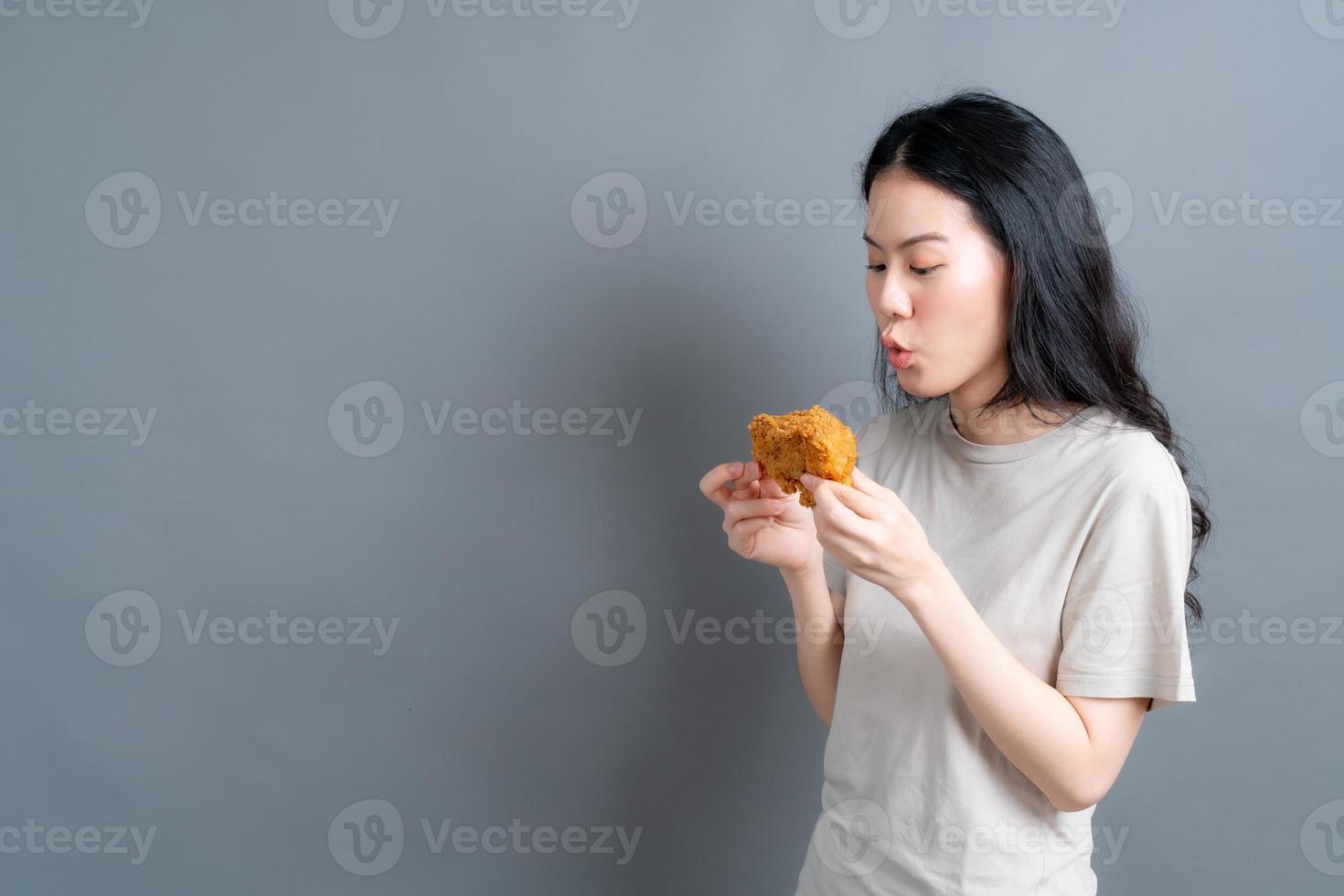 junge asiatische frau trägt einen pullover mit einem glücklichen gesicht und genießt es, gebratenes hähnchen auf grauem hintergrund zu essen foto