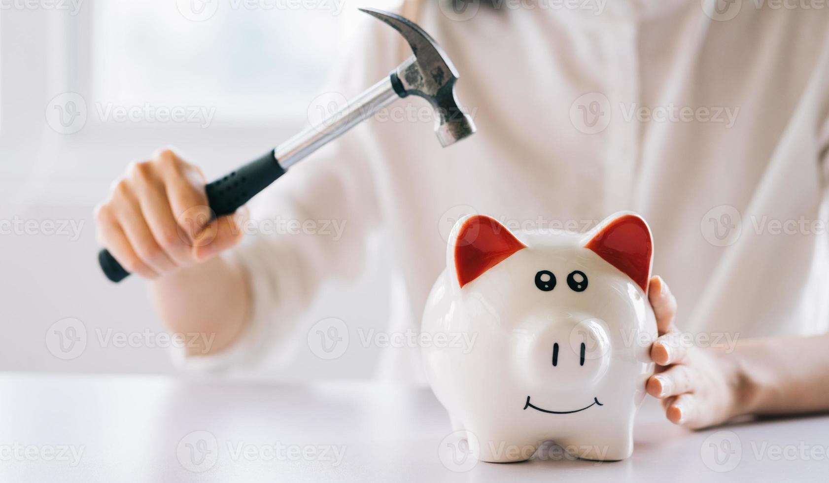 Frauenhand, die einen Hammer hält, um das Sparschwein auf dem Tisch zu zerbrechen, Geld und finanzielle Investitionen zu sparen foto