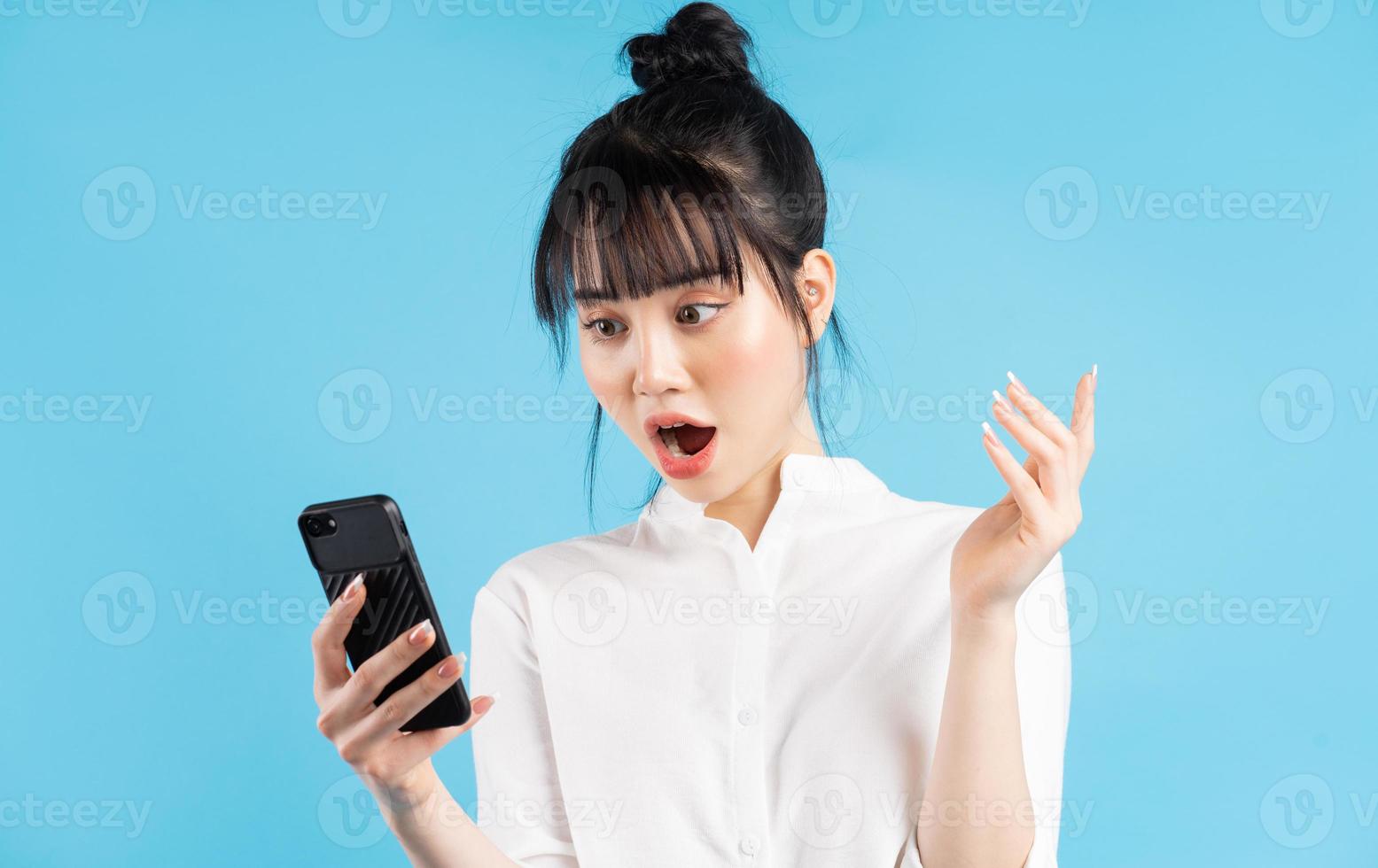 schöne asiatische frau, die telefon über blauem hintergrund mit überraschtem ausdruck hält foto