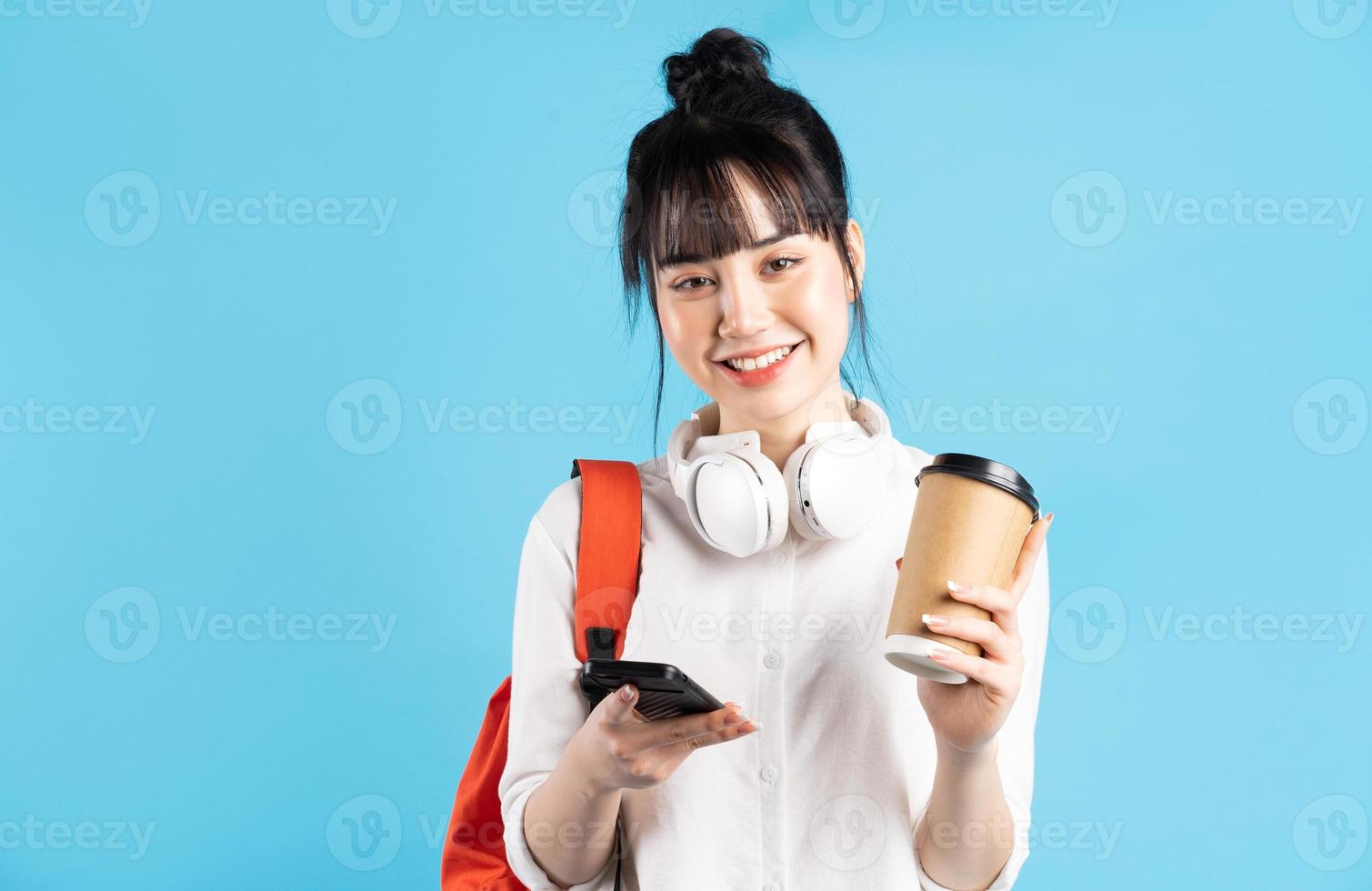 Asiatische Studentin, die Rucksack hinter dem Rücken trägt, Smartphone hält, Hals mit drahtlosen Kopfhörern, Pappbecher hält holding foto