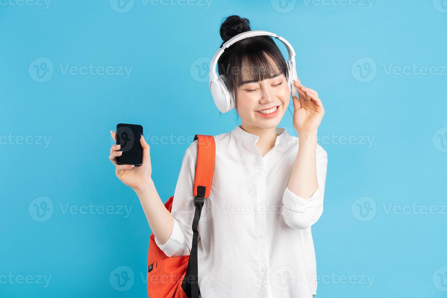 Asiatische Studentin, die Rucksack hinter dem Rücken trägt, Smartphone hält, Hals mit drahtlosen Kopfhörern, Pappbecher hält holding foto