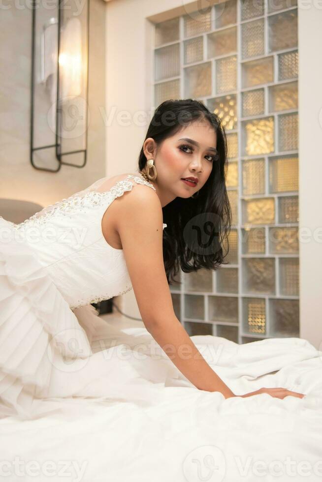 ein asiatisch Frau im ein Weiß Kleid ist posieren sexy mit ein Gesicht Das sieht aus geil auf ein Weiß Bett foto